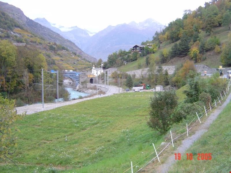 The image shows a scenic mountain landscape with a small town or village nestled in the valley. There are houses, buildings, and a road winding through the area. The landscape is surrounded by mountains and forests, with some trees in autumn colors. There