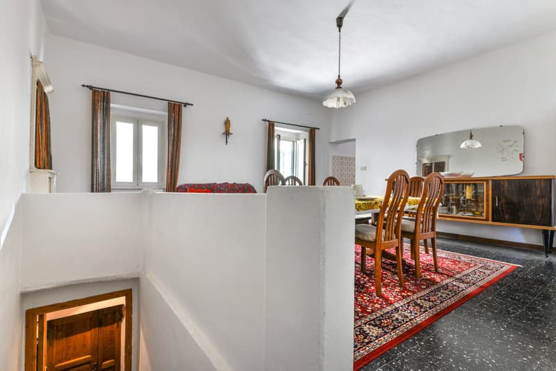 The image shows a combined living room and dining room area. The living room has a couch with colorful patterned cushions, and a fireplace. The dining room has a wooden table with chairs, and a large mirror on the wall. The room has white walls, wooden fl