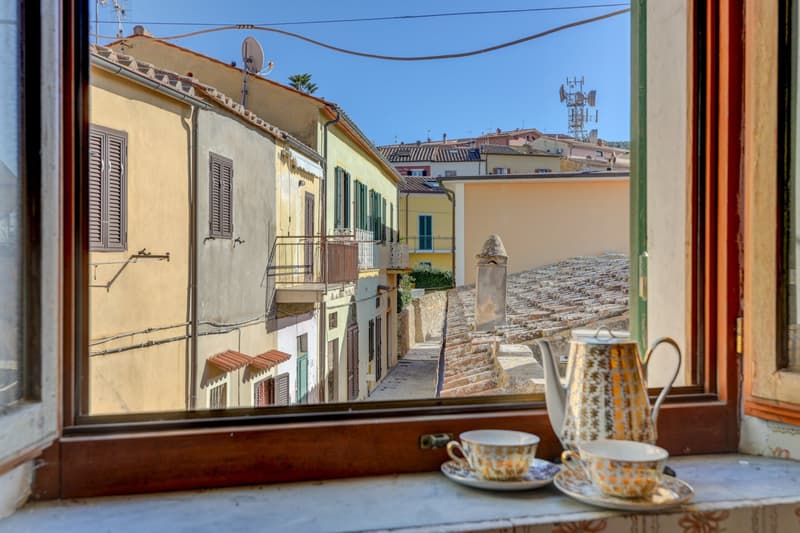 View of a quaint Italian town with colorful buildings, narrow streets, and a balcony overlooking the scene. There are some dishes and cups on the balcony, suggesting it may be a residential or cafe setting.