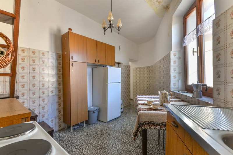 Vintage-style kitchen with wooden cabinets, refrigerator, dining table, patterned tile walls, and a chandelier overhead.