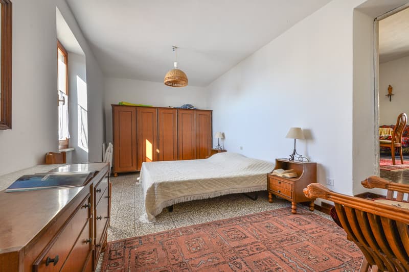 Spacious bedroom with wooden furniture, including a large wardrobe, nightstand, and bed. The room has a tiled floor and a patterned rug. There is a window and a washing machine visible in the room.