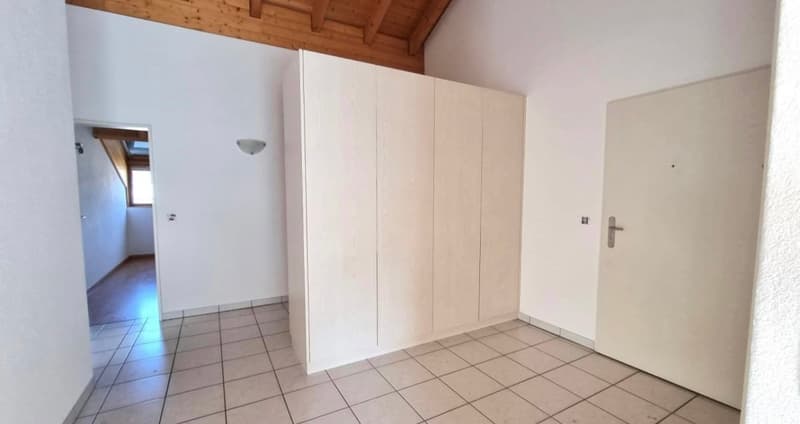 The image shows a bedroom with a tiled floor, white walls, and a wooden ceiling. There is a large built-in wardrobe along one wall, and a doorway leading to another room.