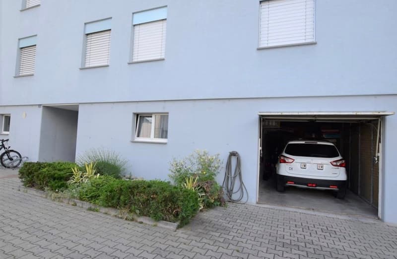 2-story white building with multiple windows, garage door open with a car parked inside, landscaped garden area in front of the building