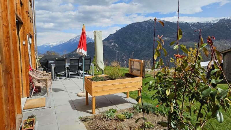 wooden planter, outdoor dining table, chairs, umbrella, wooden house, grassy area, plants, mountains in the background