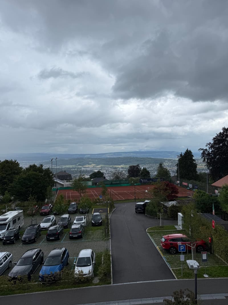 Aerial view of a parking lot with multiple cars parked, tennis courts, and a scenic view of the city in the distance.