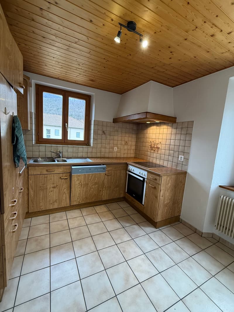 A kitchen with wooden cabinets, a sink, a faucet, a dishwasher, an oven, tiled floor, ceiling lights, a window, and a radiator.