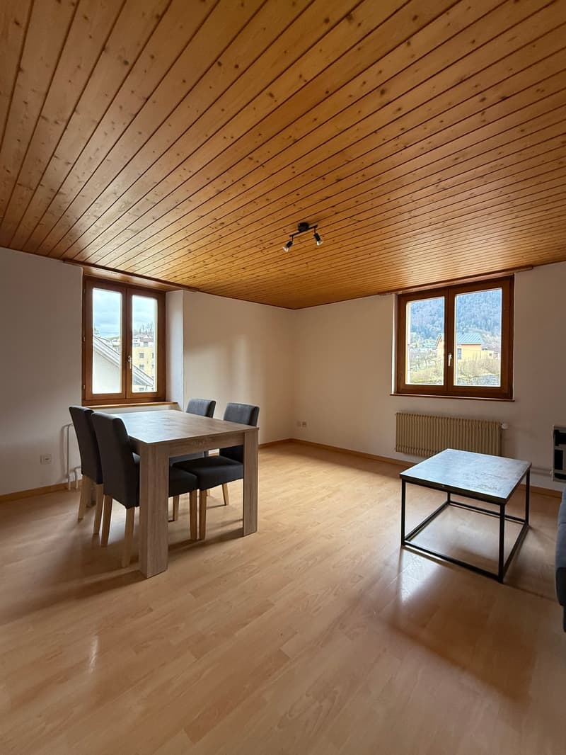 Spacious room with wooden floor, large windows, dining table, chairs, and a coffee table. A radiator is mounted on the wall.