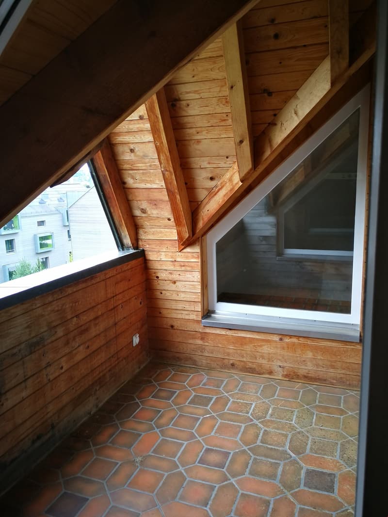 An empty attic room with wooden walls and floor, featuring hexagonal tiles and two triangular windows.