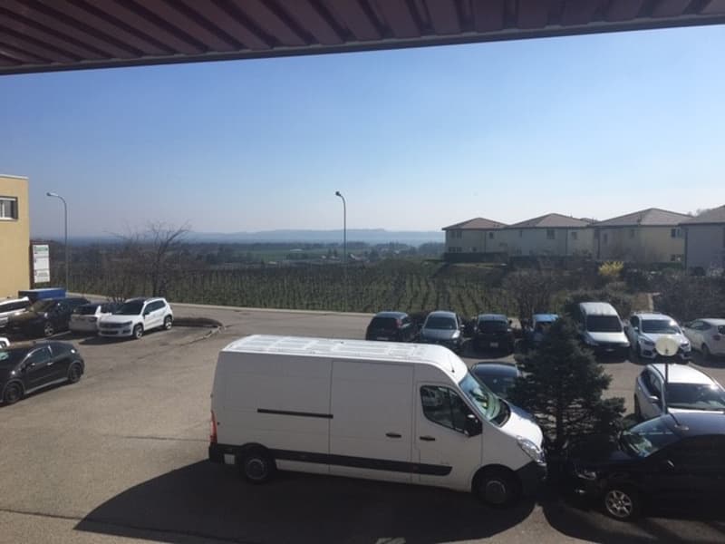 The image shows an outdoor view from a building or apartment. There are several cars parked in the foreground, and in the background, there is a hilly landscape with buildings and trees visible.