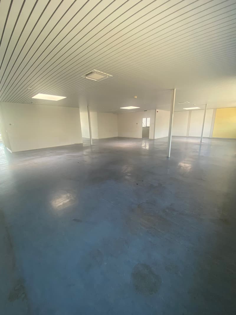 Empty warehouse space, white walls, grey floor, metal beams on ceiling, yellow accent wall, doors on far end, and stairs nearby.