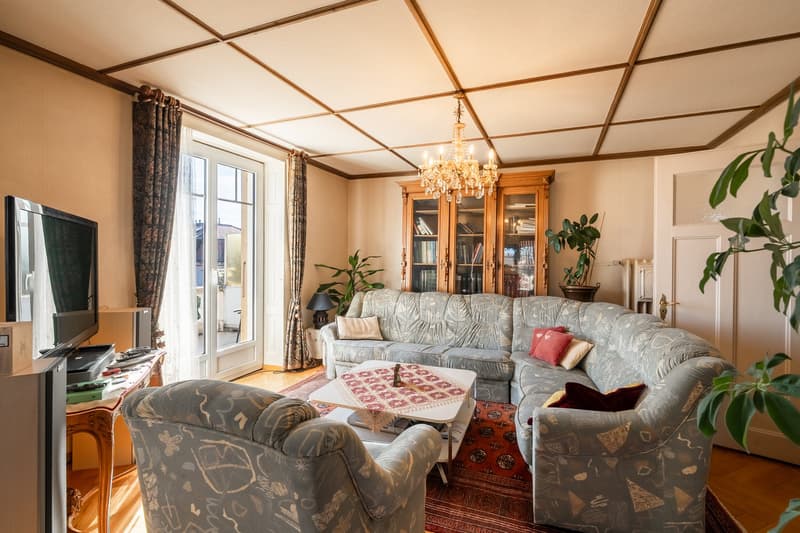 Large grey couch, wooden table, television, bookshelf, two armchairs, carpet, plants, chandelier, balcony