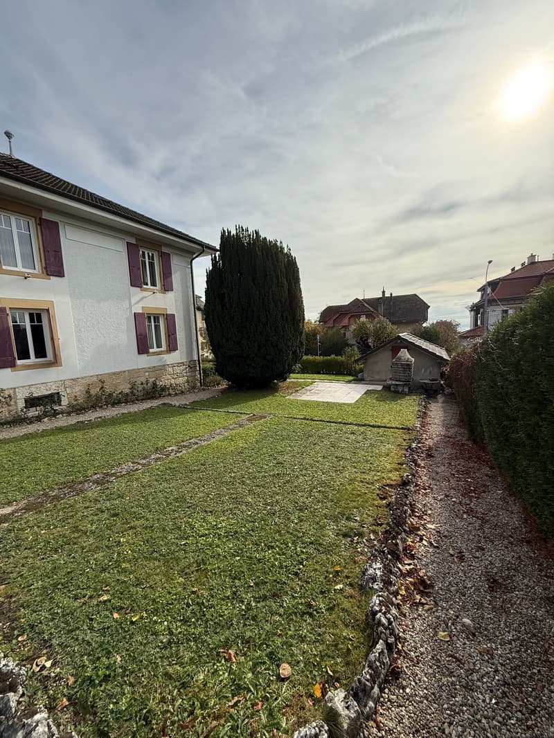 white brick house, lawn, large green bush, gravel pathway, paved area