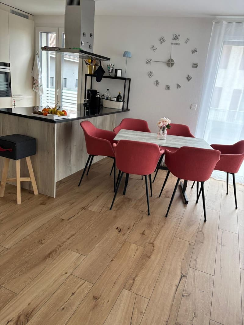 modern dining room, wooden floor, island countertop, wooden bar stools, dining table with red chairs, large wall clock, white cabinets, kitchen appliances