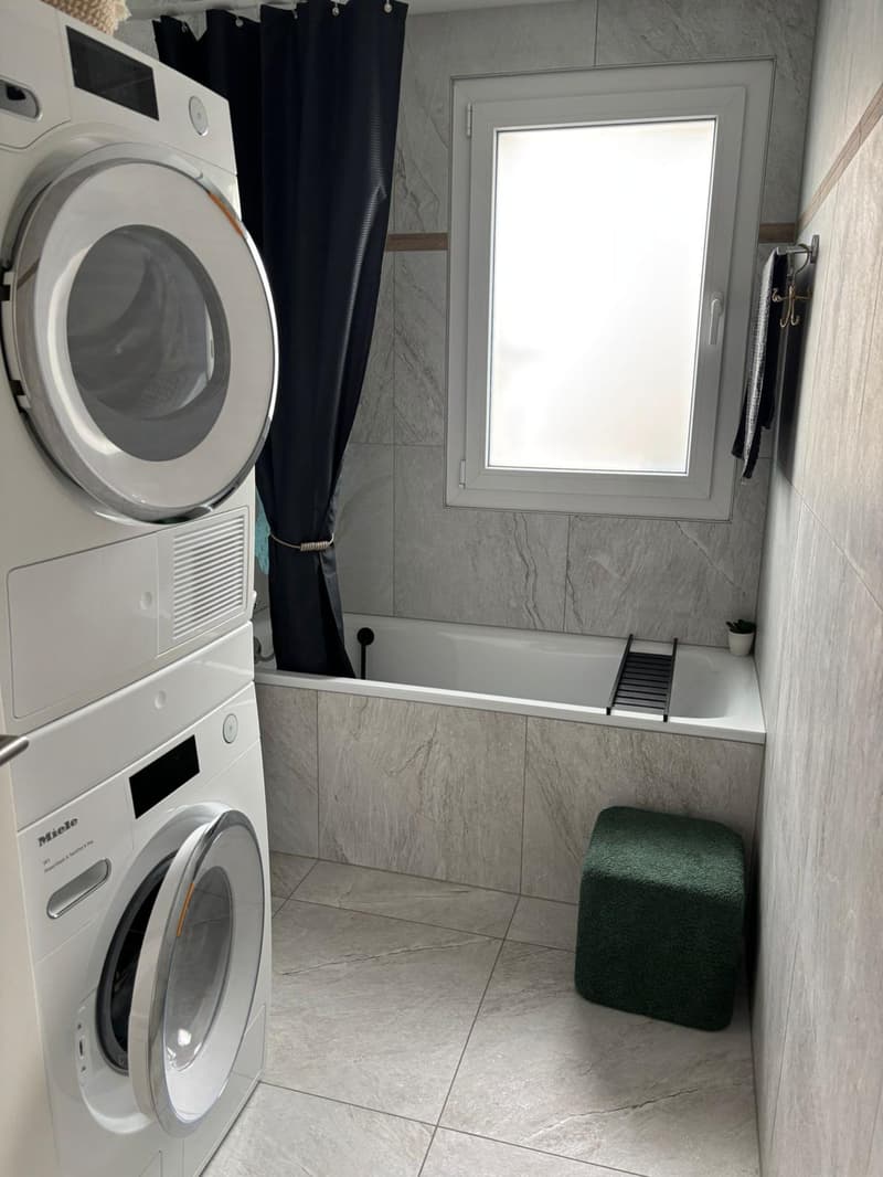 Modern white tiled bathroom with washing machine, bathtub, window, and small ottoman