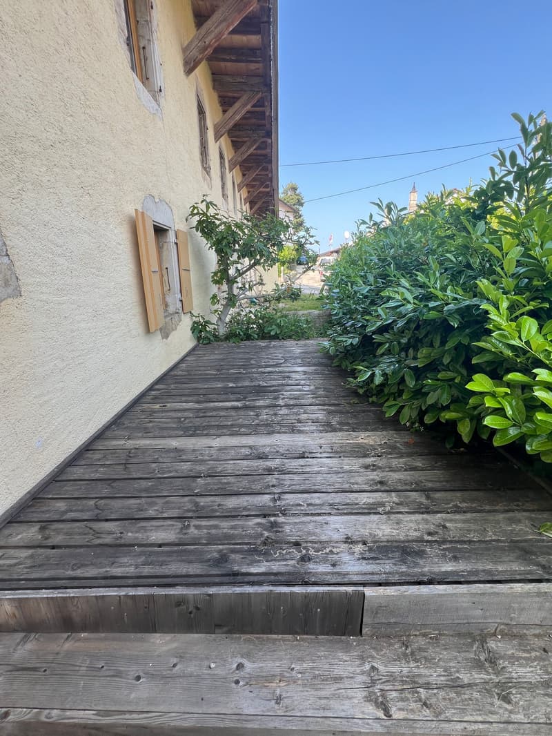 Wooden walkway leading along the side of an old building with a tiled roof. The walkway is surrounded by lush greenery, including shrubs and plants. The building has a rustic, weathered appearance with wooden beams and a stucco exterior.