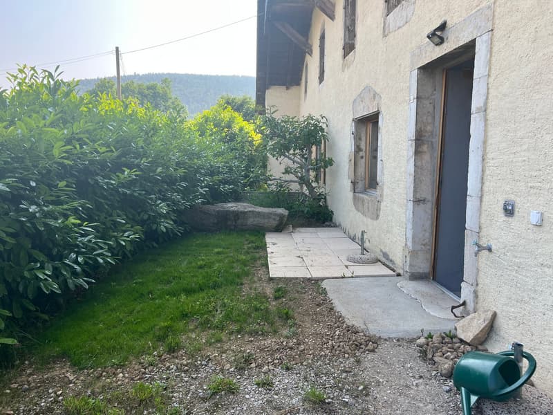 Old stone building with a small garden area, grassy lawn, and lush vegetation surrounding the property. There is a concrete walkway leading to the entrance of the building.