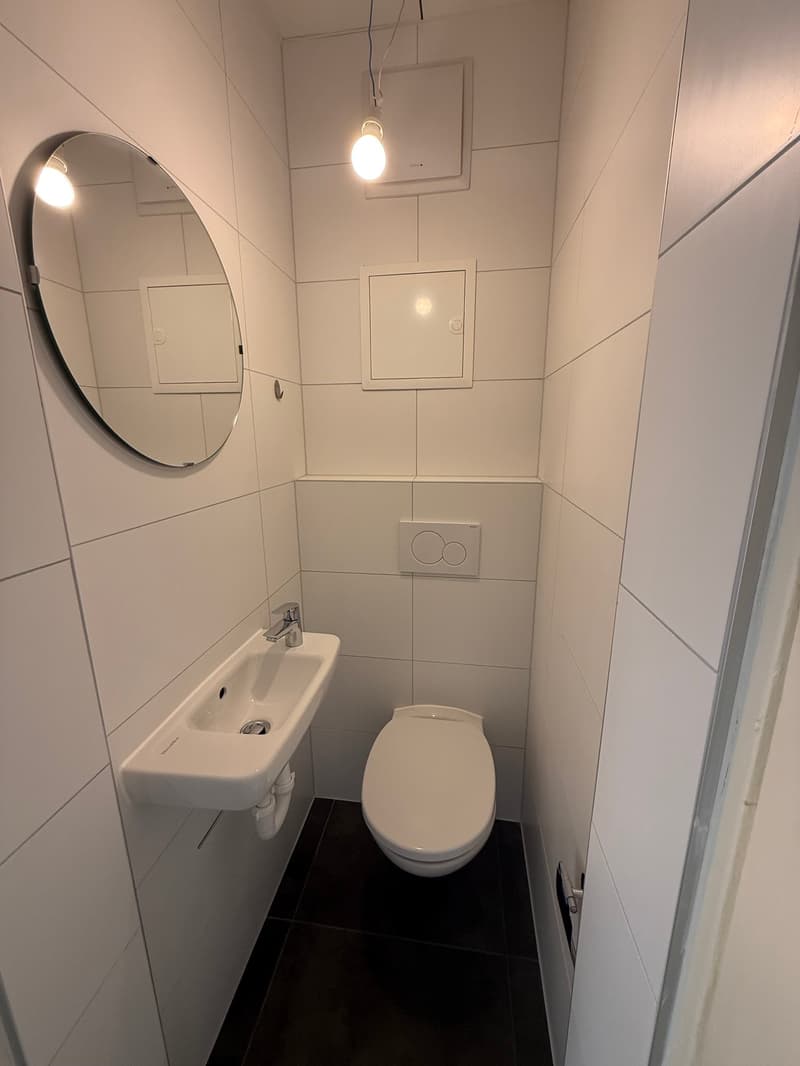 small bathroom with white tiles, white sink, white toilet, round mirror, ceiling light
