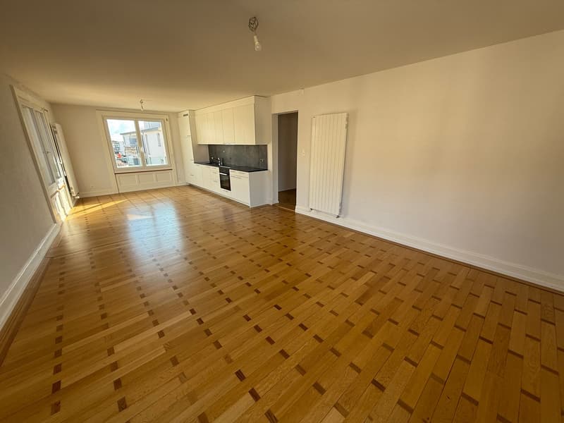 Empty room with wooden floor, white walls, kitchen with white cabinets, black countertops, and a window.