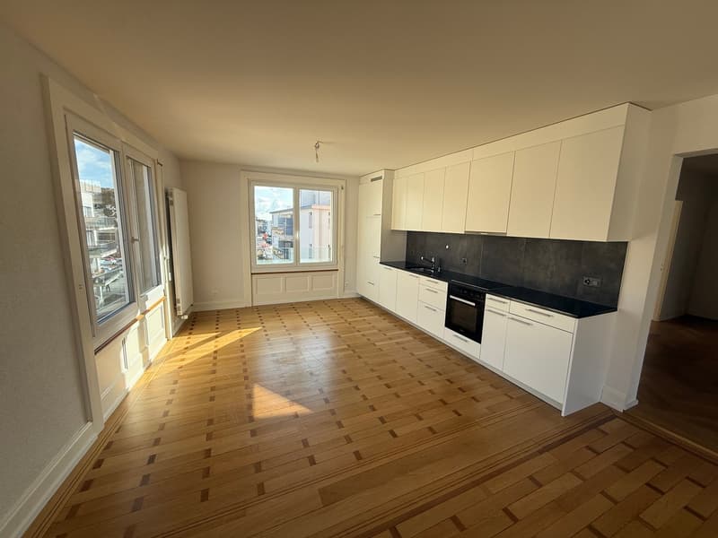 Empty kitchen with wooden floor, white cabinets, black countertops, built-in oven, sink, and faucet, large windows