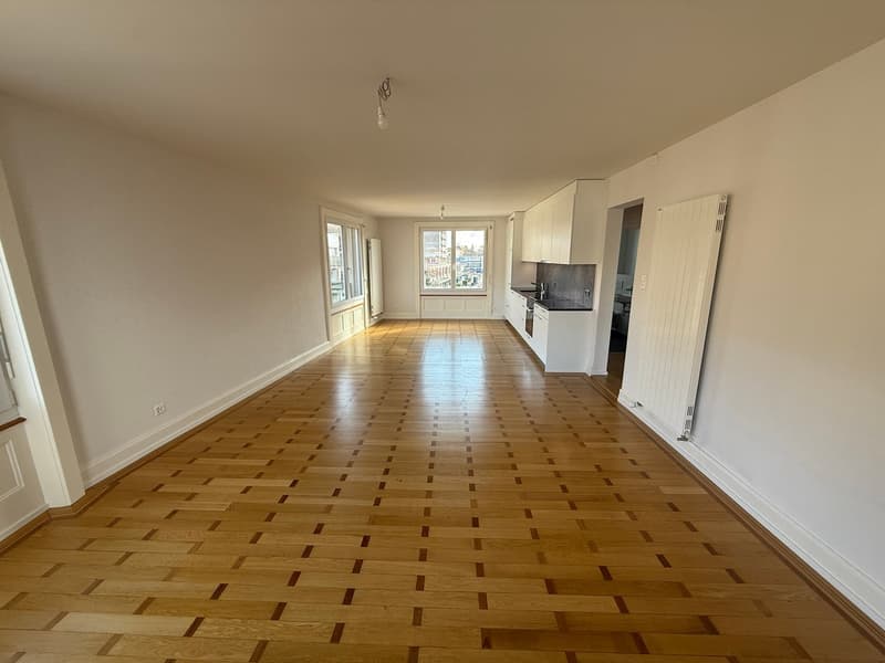 Empty room with wooden floor, white walls, windows, kitchen area, and a hallway leading to other rooms.