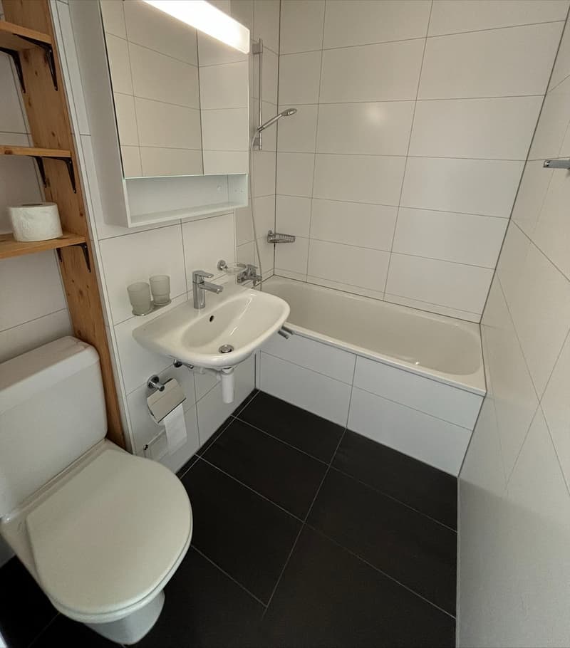 Bathroom with white tiles, white toilet, white sink, white bathtub, wooden shelf with toilet paper, shower head, and faucet