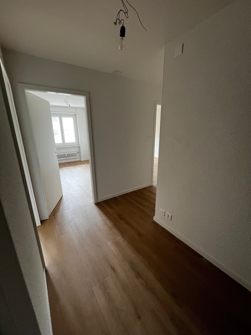 Empty room with wooden floor, white walls, open door, windows, power outlets, and a light fixture