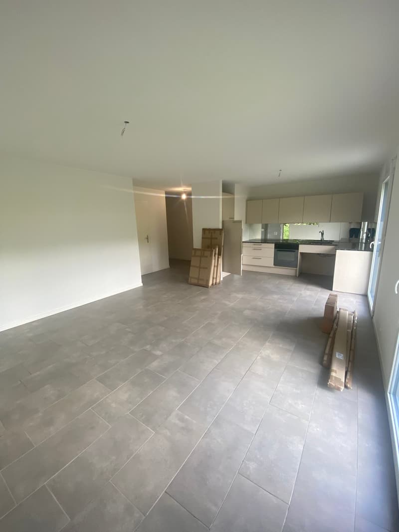 Empty room with gray tiles, white walls, kitchen area with cabinets, sink, and oven, glass door leading to another room, boxes on the floor.