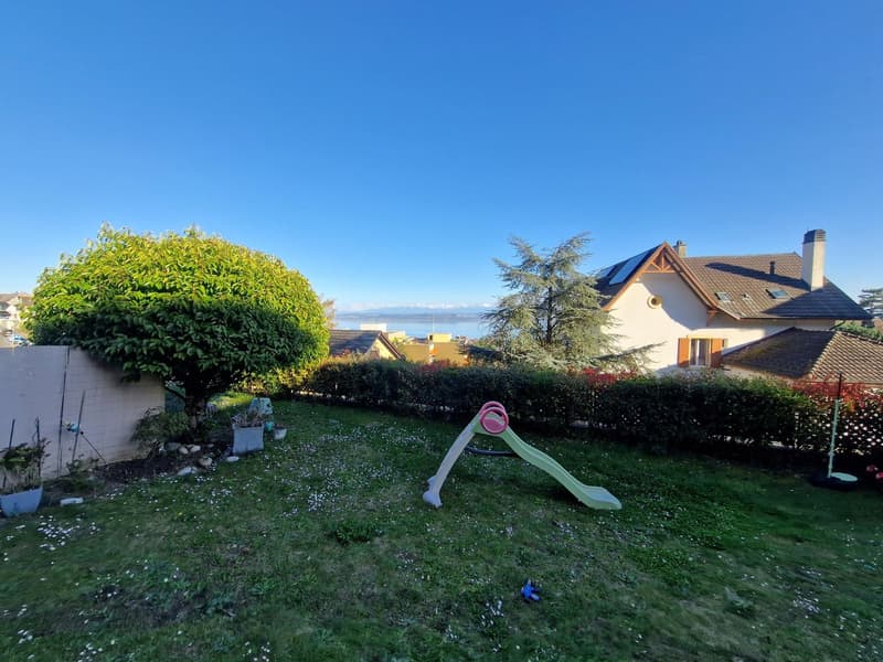 A garden with a slide, a tree, and a house in the background with a view of the lake.