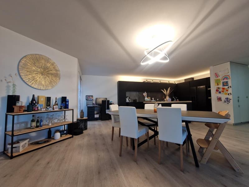 Dining area with a large table, white chairs, wooden floor, modern ceiling light, a shelf with bottles, a round wall decoration, and a kitchen area with black cabinets and a refrigerator.