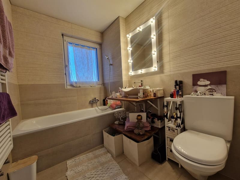 Bathroom with bathtub, mirror with lights, toilet, shelves, towels, and a window.