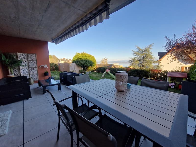 Outdoor dining area with a table and chairs, a sofa, a coffee table, a potted plant, a vase, a rug, a canopy, a garden, a house, a tree, a lake, and a mountain in the distance.