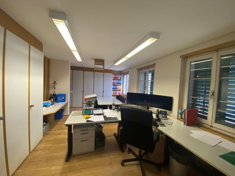 Two desks with office equipment and chairs, shelves, large windows, wooden floor.
