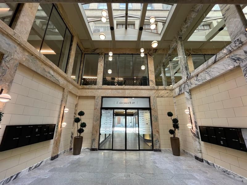 Marble and white tiled building with multiple floors and a large entrance with glass doors, glass walls, potted plants, and hanging lamps