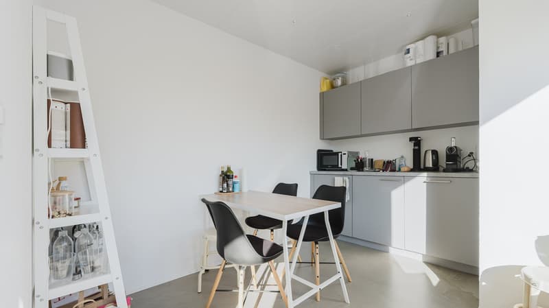 Modern kitchen with gray cabinets and white walls, a table with stools, kitchen appliances, and a bookshelf.