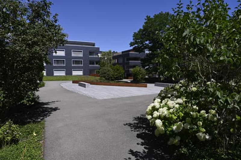 Modern apartment building with gray exterior, concrete driveway, greenery, and benches