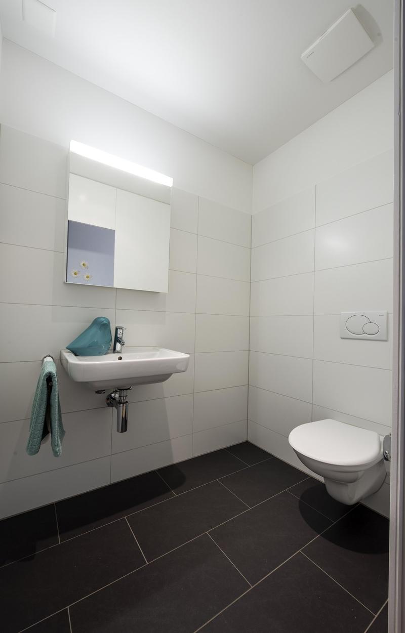 Bathroom with white tiles, white sink, mirror, white toilet, green towel, black floor