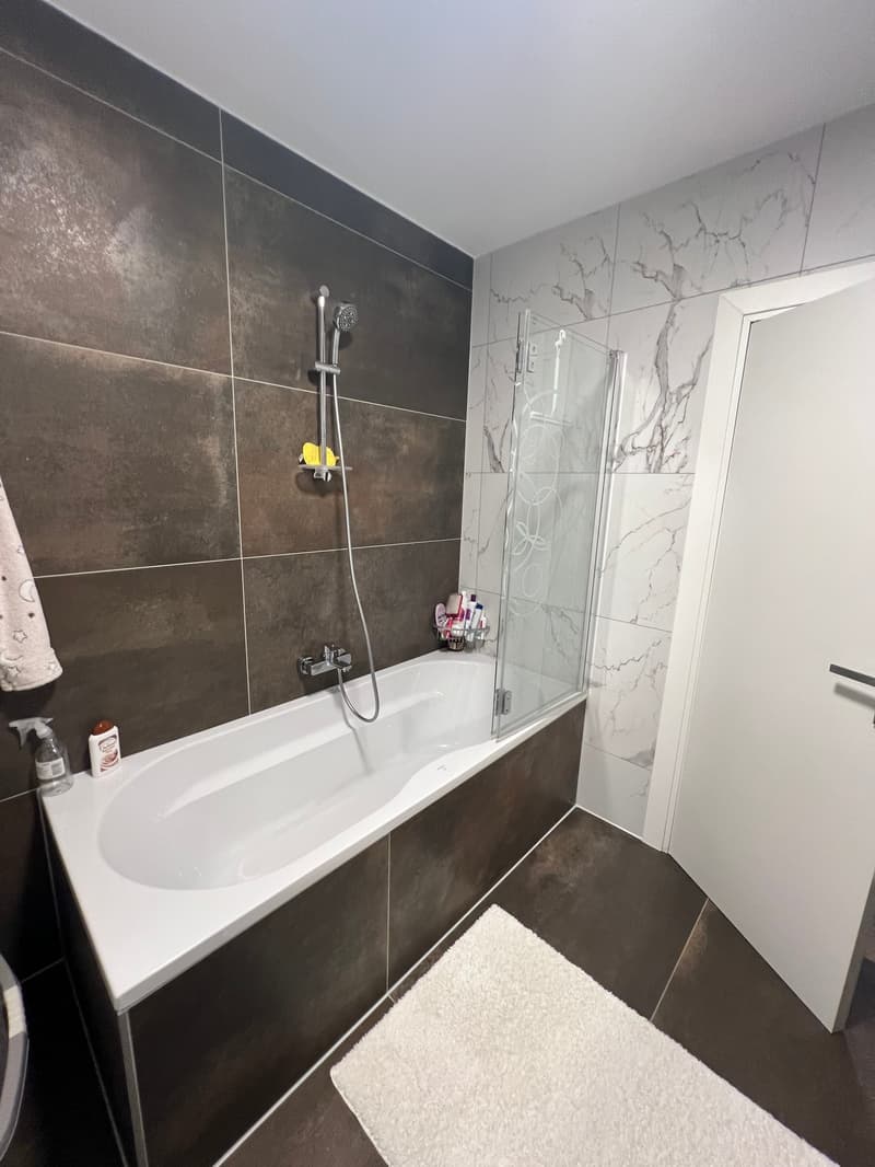 Bathtub, shower, white tiled walls, black tiles, white towel, white rug, white door, toiletries