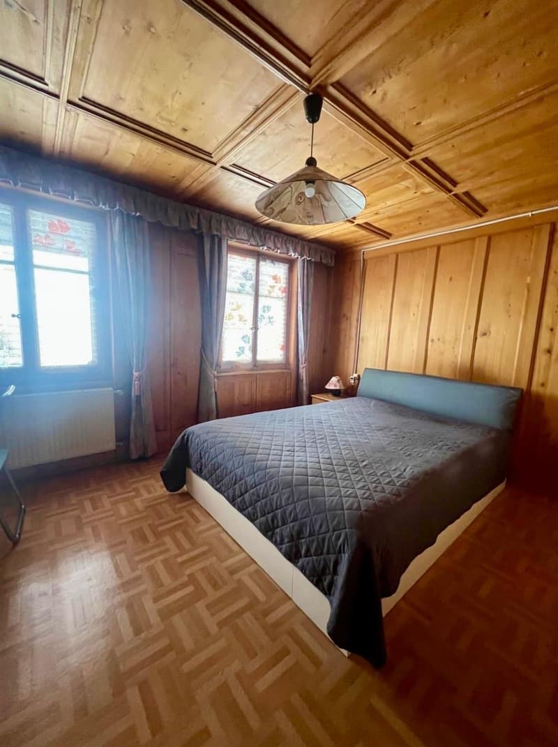Large bedroom with wooden bed, bedside table, white radiator, wooden floor, large windows with curtains, pendant lamp, wooden wall panels