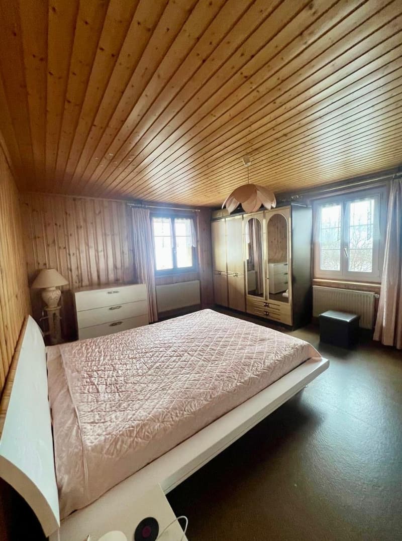 Large bedroom with double bed, wooden walls, wooden floor, white wardrobe, large windows, lamp, and radiator