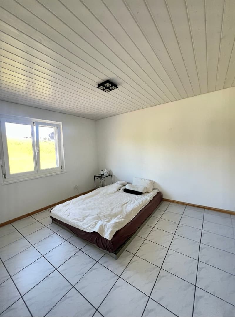 Empty room with white walls, white tiled floor, a bed with white sheets and pillows, a small table with a candle, and a window with a view of a field.