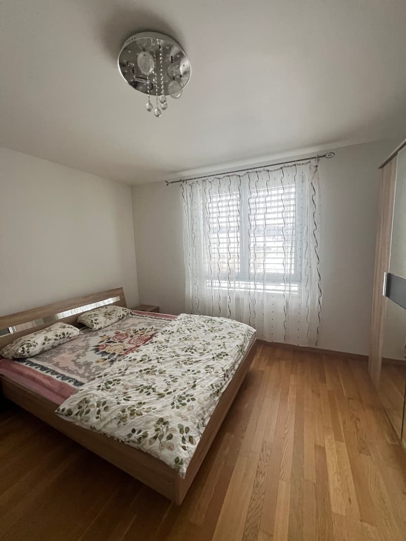 Bedroom with wooden bed, floral bedspread, two pillows, wooden nightstand, white curtains, wooden floor, chandelier