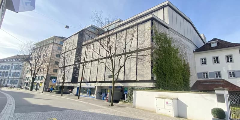 Modern building with white and black exterior, glass windows, parking sign, trees, and a small garden