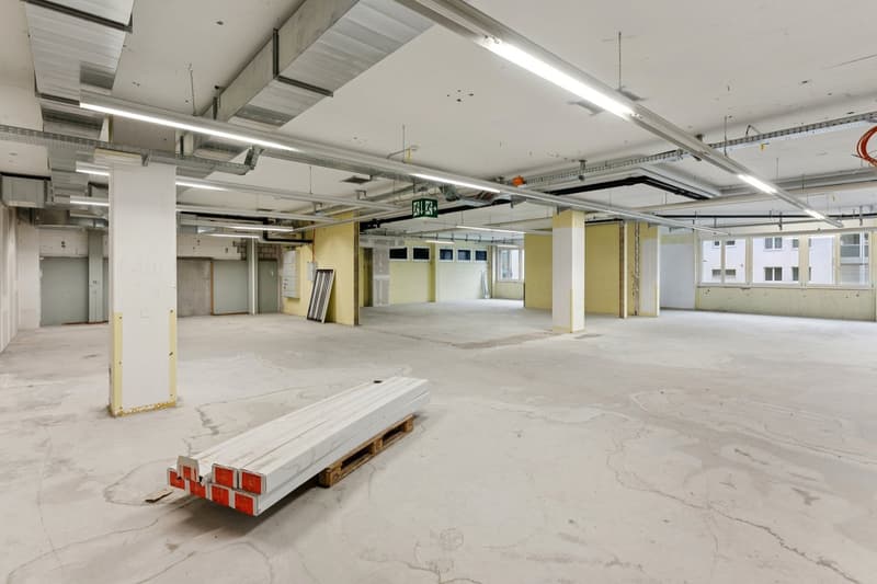 Empty room with exposed ceiling beams and fluorescent lighting, concrete floor, no furniture.