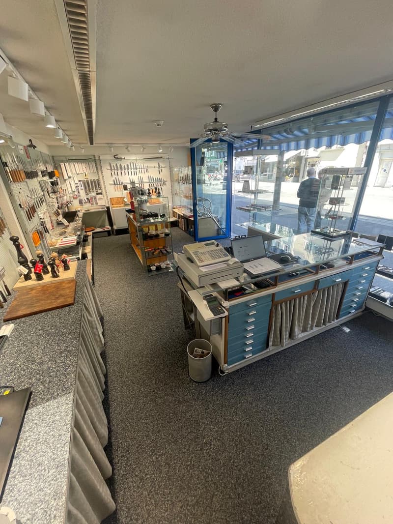 Interior of a store with a reception desk, glass display cases, and various items on display. The store has a modern design with gray carpet, white walls, and ceiling lights. There are glass windows with blue awnings, and a person is standing outside.