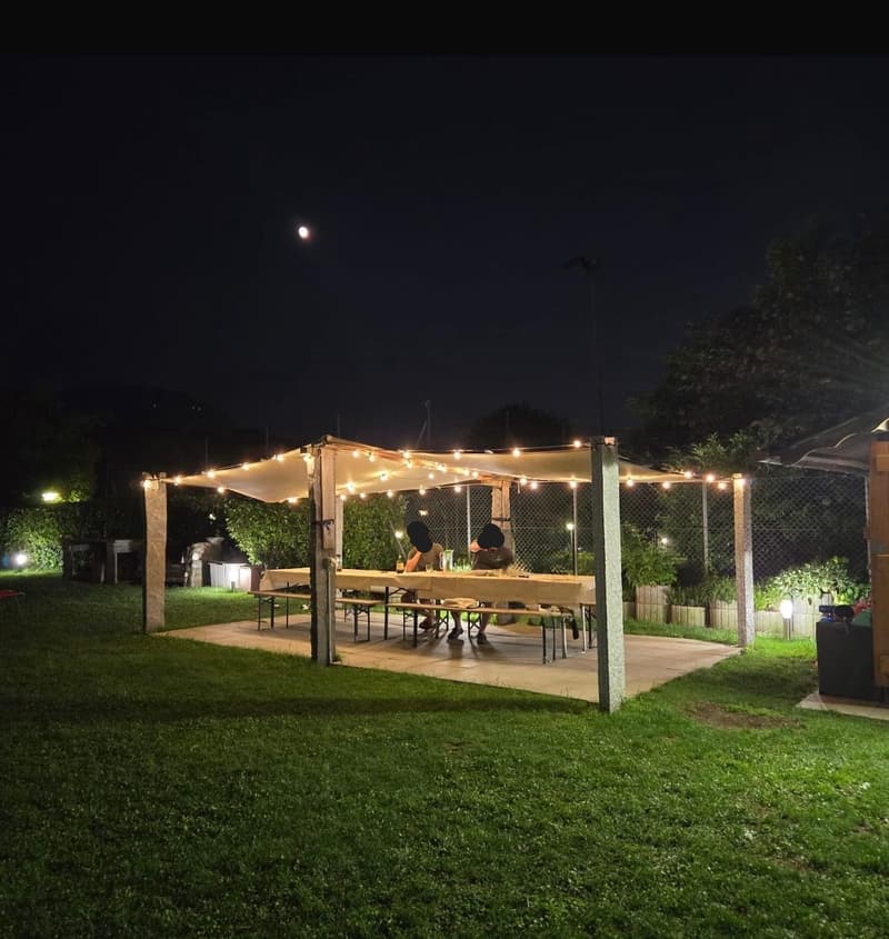 Covered outdoor seating area with string lights, wooden benches, and a fireplace. The area is surrounded by greenery and located in a backyard setting.