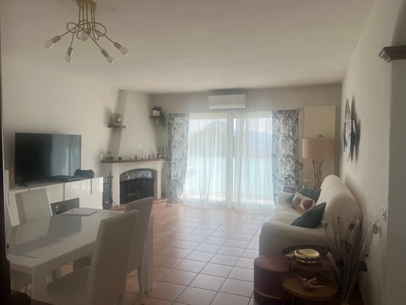 Spacious living room with tile flooring, large windows providing natural light, a fireplace, and a balcony or terrace. The room is furnished with a sofa, a dining table, and a TV stand.