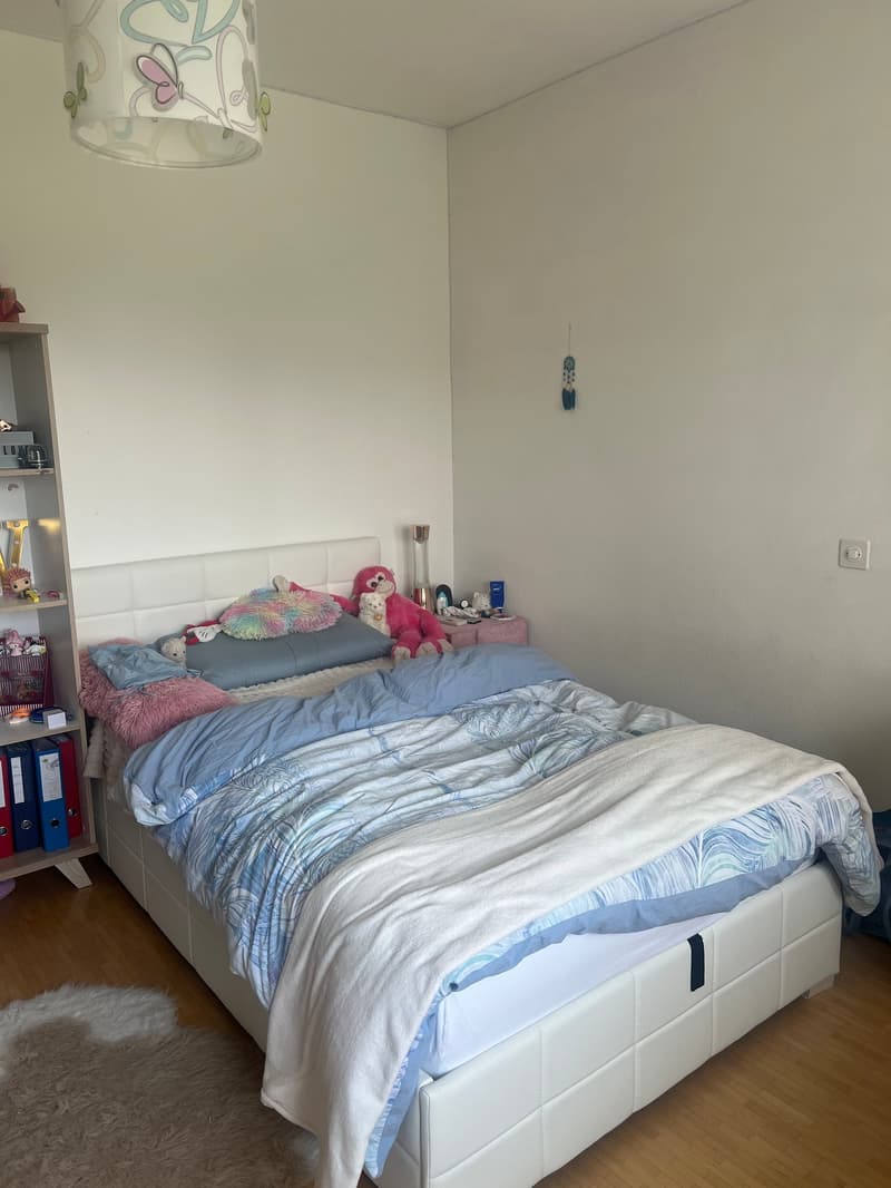 Bedroom with a double bed, colorful bedding, and plush toys. There is a shelving unit and a decorative lamp shade visible.