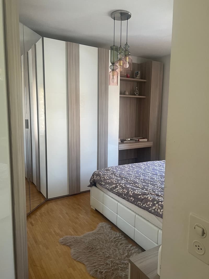 Bedroom with a large wardrobe, a bed with a patterned bedspread, a shelf unit, and a pendant light fixture with colored glass shades.