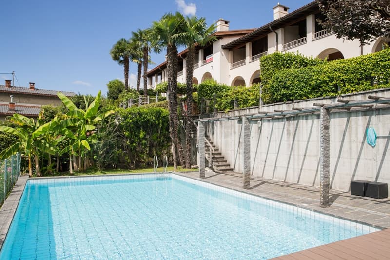 Two-story Mediterranean-style building with arched balconies, surrounded by palm trees and lush greenery. There is a large swimming pool in the foreground with a wooden deck area.