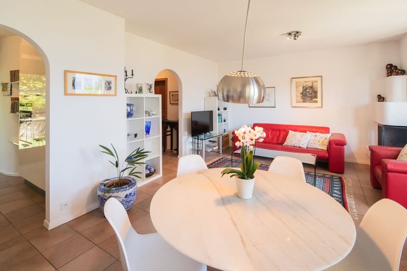 Open-concept living room with white walls, hardwood floors, a round dining table, red sofa, TV stand, and a hanging light fixture. There is a plant and artwork on the walls.
